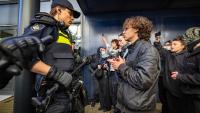 Een politieagent staat tegenover een groep mensen van wie er één tegen de agent praat. De agent houdt een wapenstok en een radio vast. Verschillende omstanders op de achtergrond observeren het tafereel en nemen het op met hun telefoon.