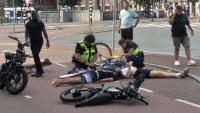 Twee politieagenten verlenen hulp aan een gewonde die op straat ligt, vlakbij omgevallen fietsen en een elektrische fiets. Er zijn omstanders aanwezig en het incident lijkt zich op een kruispunt te hebben voorgedaan.