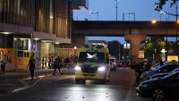 In de schemering staat een ambulance met brandende koplampen geparkeerd in een straat in de stad, vlakbij een metrostation. Er zijn politieagenten en omstanders in de buurt en het gebied is met afzetlint afgezet. Er zijn verschillende geparkeerde auto’s e